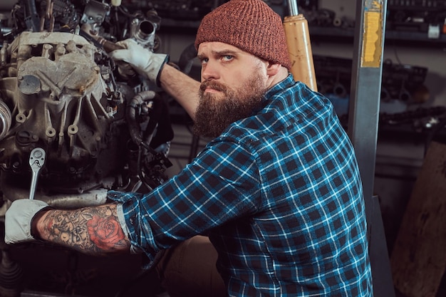 Kostenloses Foto brutaler tätowierter bärtiger mechaniker repariert den automotor, der auf der hydraulischen hebebühne in der garage angehoben wird. tankstelle.