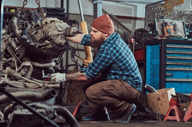 Brutaler tätowierter bärtiger Mechaniker repariert den Automotor, der auf der hydraulischen Hebebühne in der Garage angehoben wird. Tankstelle.