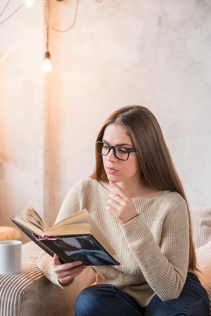 Buch der jungen Frau ernst beim Sitzen auf Sofa