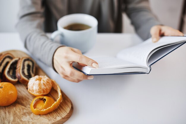 Buch ist wie eine Kammer des Wissens. Intelligente moderne Frau liest Lieblingsroman während des Frühstücks, genießt heißen Tee in ruhiger und gemütlicher Atmosphäre, schält Mandarine und isst