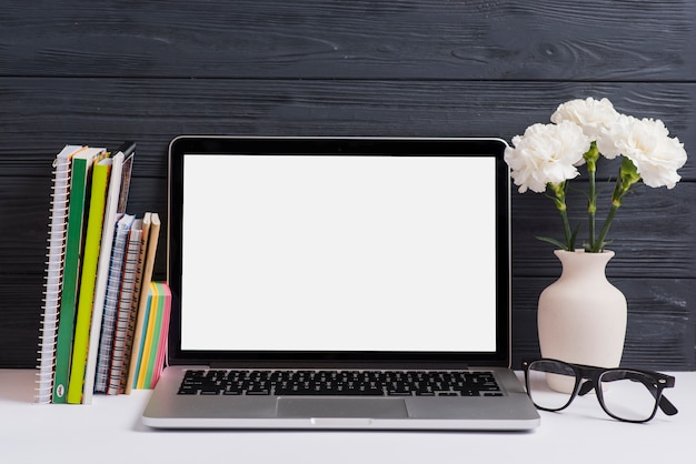 Bücher; Haftnotizen; Laptop; Brille und Vase auf weißem Schreibtisch gegen Holzwand