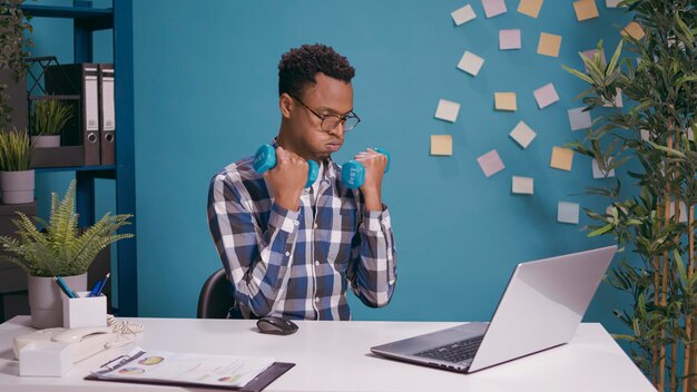 Büroangestellter trainiert mit Hanteln, um Muskeln zu trainieren, während er am Schreibtisch einen Laptop benutzt. Mitarbeiter heben Gewichte und machen körperliche Aktivität mit Fitnessübungen, arbeiten am Computer für Unternehmen.