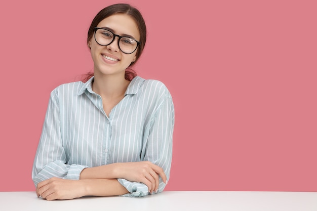 Büroarbeit, job, beruf, beruf, karriere und erfolgskonzept. schöner fröhlicher junger weiblicher manager in der stilvollen brille und im formellen hemd, die arme auf dem schreibtisch verschränkt halten und selbstbewusst lächeln