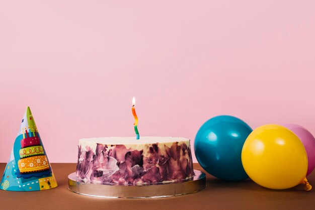 Bunte belichtete Kerze auf Geburtstagskuchen mit Partyhut und -ballonen auf Schreibtisch gegen rosa Hintergrund