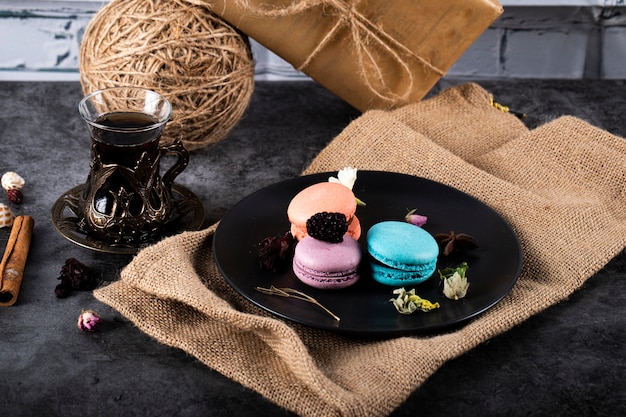 Bunte Macarons in einer schwarzen Untertasse und einem Glas Tee auf einem schwarzen Tisch