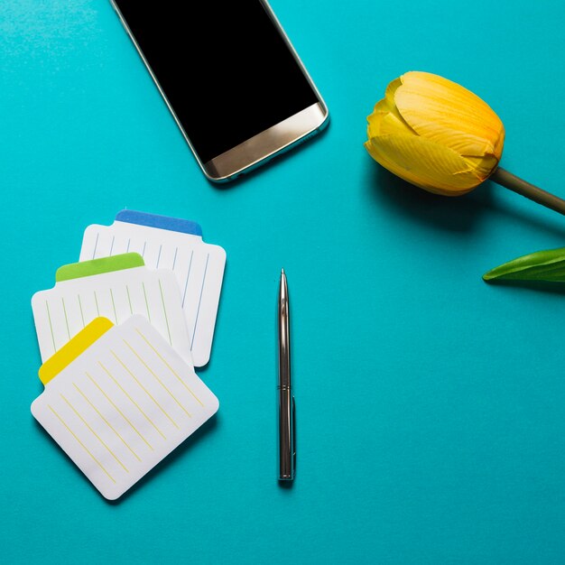 Bunte Notizblöcke mit Stift; Handy und gelbe Tulpen auf blauem Hintergrund