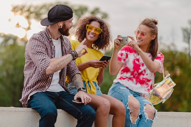 Bunte stilvolle glückliche junge Gesellschaft der Freunde, die Park sitzen