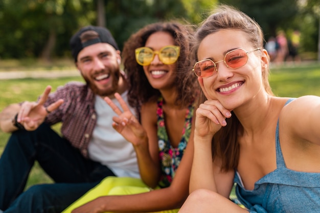 Bunte stilvolle glückliche junge Gesellschaft von Freunden, die Park, Mann und Frauen sitzen, die Spaß zusammen haben