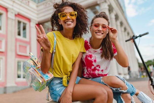 Bunte stilvolle glückliche junge Mädchenfreunde lächelnd sitzen in der Straße, Frauen, die Spaß zusammen haben