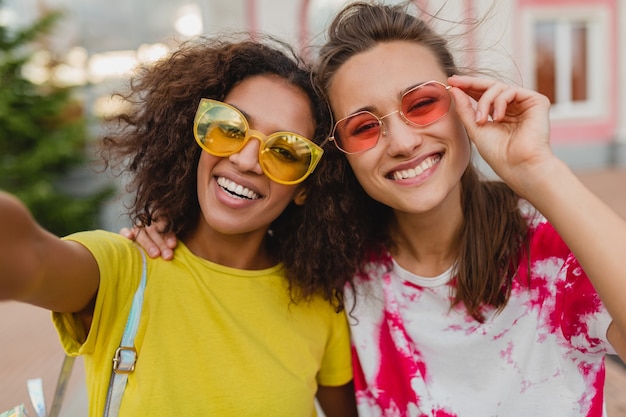 Buntes Porträt von glücklichen jungen Mädchenfreunden, die lächelnd sitzen auf der Straße Selfie-Foto auf Handy, Frauen, die Spaß zusammen haben