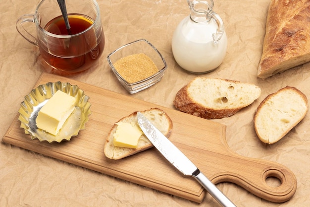 Butter, brotscheibe und messer auf schneidebrett. tee, milch und französisches baguette auf dem tisch. hintergrund aus braunem papier. ansicht von oben