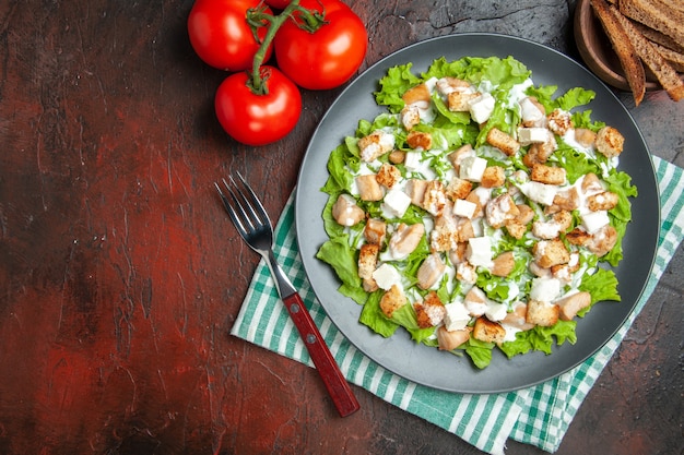 Cäsarsalat von oben auf ovaler Platte grün-weiß karierte Servietten-Tomatengabel auf dunkelrotem Hintergrund