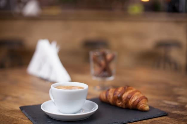 Cappuccino-Kaffee in einer weißen Tasse auf einem Holztisch neben einem köstlichen Croissant. Leckere Schlange. Vintage-Café.