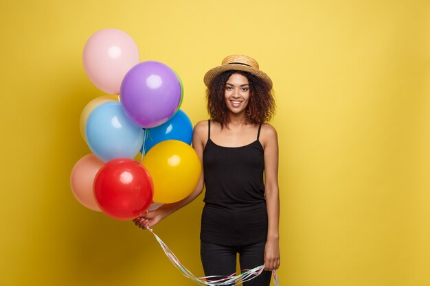 Celebration Concept - Close up Portrait glücklich junge schöne afrikanische Frau im schwarzen T-Shirt lächelnd mit bunten Party Ballon. Gelbe Pastellstudio Hintergrund.