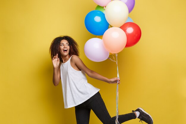 Celebration Concept - Close up Portrait glücklich junge schöne afrikanische Frau im schwarzen T-Shirt lächelnd mit bunten Party Ballon. Gelbe Pastellstudio Hintergrund.