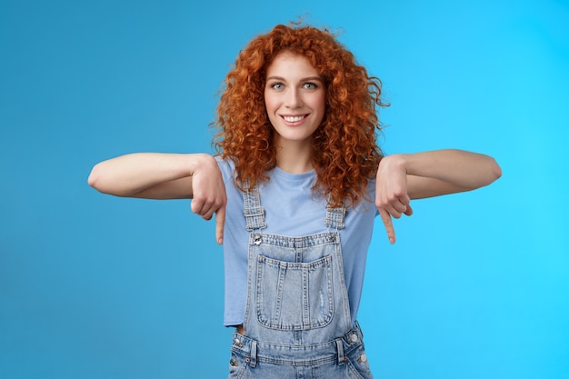 Charismatische, freche, kokette Rothaarige, die gewagtes Ingwermädchen lockiger Haarschnitt zeigt, der nach unten zeigt, mit den Zeigefingern, die allgemein enthusiastisch lächeln, erkunden Sie den neuen Laden, der Promo wie coole Werbung auf blauem Hintergrund zeigt.