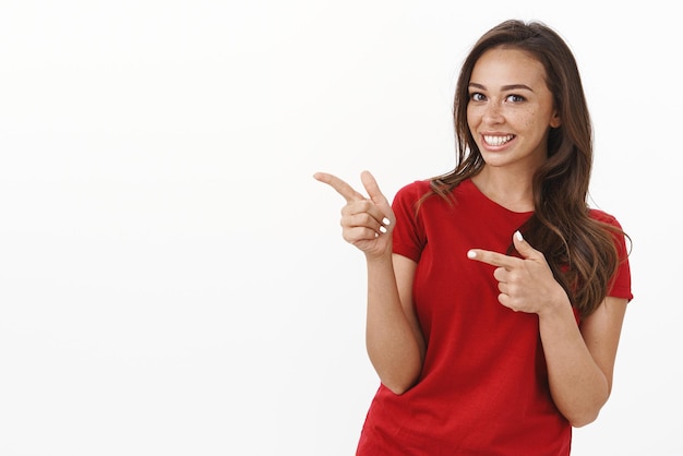 Charismatische, freundliche, brünette Frau in rotem T-Shirt mit frechen Zeigefingerpistolen, die auf einen leeren weißen Kopierraum gelassen wurden, freudig lächelnd und optimistisch, laden Sie ein, sich ein tolles Event-Promotion-Produkt anzusehen