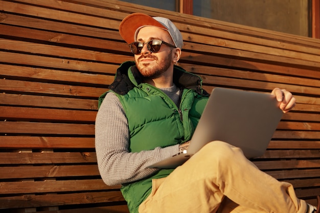 Charismatischer junger glücklicher männlicher Freiberufler mit Stoppeln, die auf Holzbank mit tragbarem Computer sitzen
