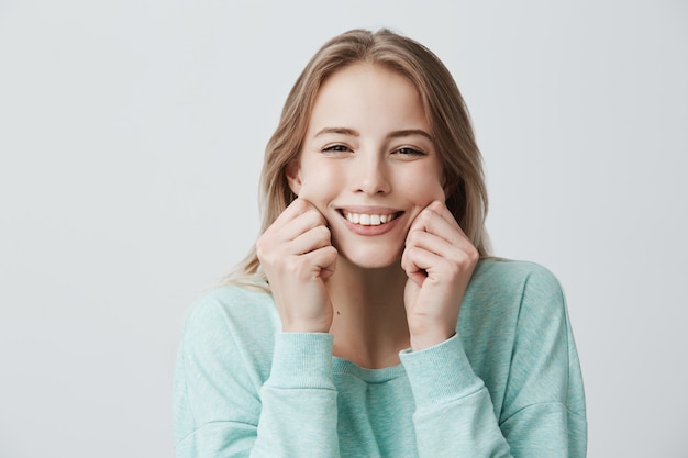 Charmant breit lächelnd mit perfekten Zähnen junge europäische Frau mit blonden langen Haaren trägt hellblauen Pullover, kneift die Wangen, verspottet, hat gute Laune und Spaß. Gesichtsausdrücke und Emotionen