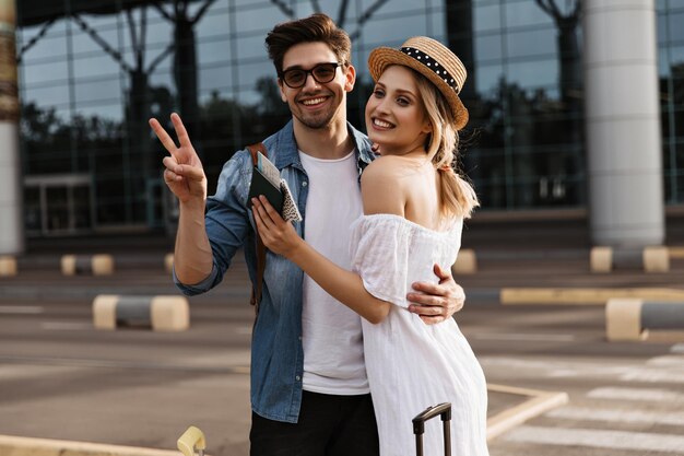 Charmante blonde Frau in hutweißem Kleid und fröhlicher brünetter Mann in Jeansjacke mit Sonnenbrille lächelt und zeigt vsign