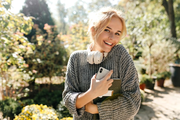 Charmante blonde grünäugige Frau in stilvollem grauem Outfit lächelnd mit Telefon und schwarzem Notizbuch Dame mit Kopfhörern um den Hals geht im Park spazieren