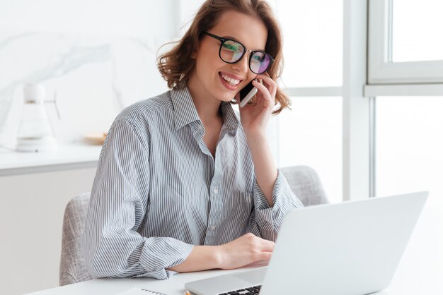 Charmante brünette Frau in Brille und Freizeitkleidung, die auf Handy spricht, während sie am Tisch sitzt und Laptop-Bildschirm betrachtet