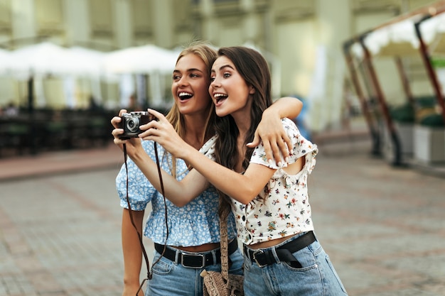 Charmante brünette und blonde Frauen in stylischen Blumenblusen und Jeanshosen lächeln und umarmen sich draußen