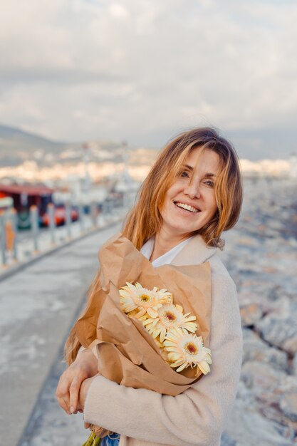 Charmante Dame, die mit Blumen am Meer während des Nachmittags lächelnd steht und freudig schaut.