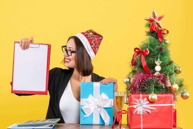 Charmante Dame im Anzug mit Weihnachtsmannhut und Brille, die Dokumente hält, die Geschenk im Büro zeigen