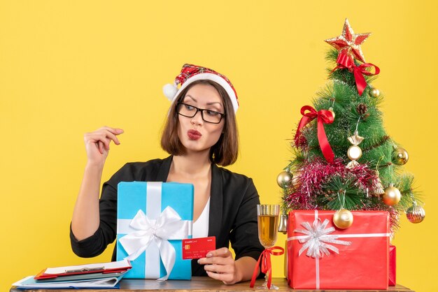 Charmante Dame im Anzug mit Weihnachtsmannhut und Brille, die Geschenk und Bankkarte zeigt, die hinten im Büro auf gelb lokalisiert zeigen