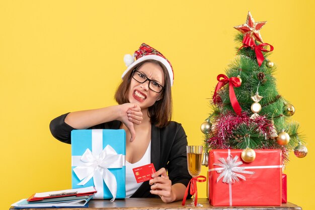 Charmante Dame im Anzug mit Weihnachtsmannhut und Brille, die Geschenk- und Bankkarte zeigt, die negative Geste im Büro auf Gelb lokalisiert macht