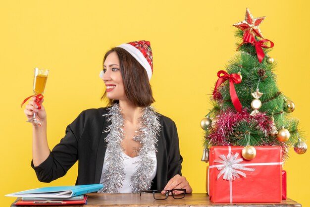 Charmante Dame im Anzug mit Weihnachtsmannhut und Neujahrsdekorationen, die Wein im Büro auf gelb lokalisiert erheben