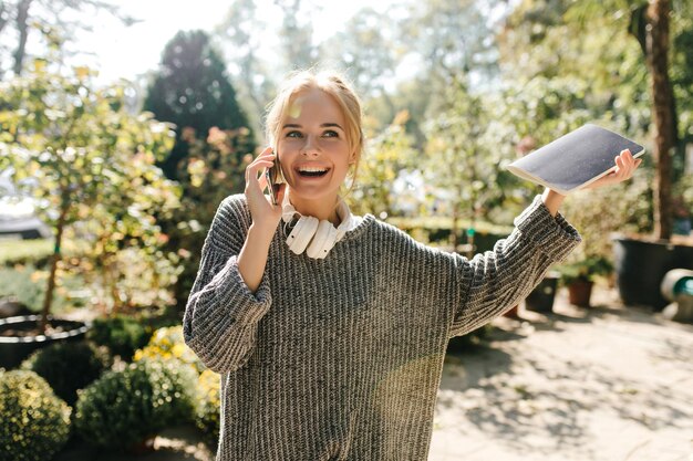 Charmante Dame in übergroßem Pullover, die glücklich telefoniert und ein Notizbuch in den Händen hält Porträt eines Mädchens mit Kopfhörern um den Hals im Garten