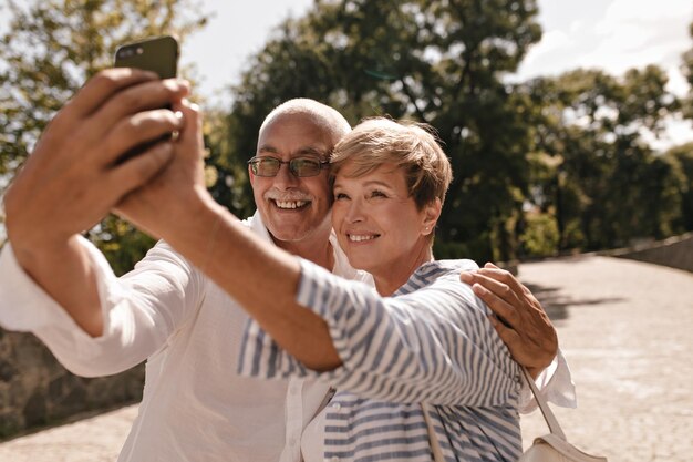 Charmante Dame mit moderner blonder Frisur in gestreifter Bluse lächelt und macht Selfie mit glücklichem Mann mit grauem Schnurrbart in Brille und Hemd im Freien