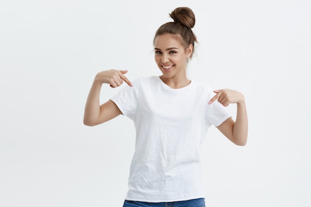 Charmante emotionale kaukasische Frau, die nach unten oder auf ihr T-Shirt zeigt, während sie freudig lächelt und positive Gefühle ausdrückt