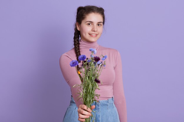 Charmante Frau hält Blumenstrauß von Feldblumen, Frau bietet Blumen zu jemandem, Mädchen mit Zöpfen, das isoliert über lila Wand, lächelndes junges Mädchen aufwirft.