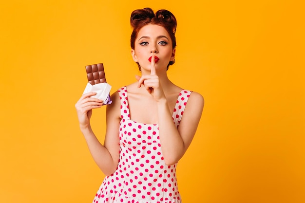 Charmante frau im polkadot-kleid, die lippen mit dem finger berührt studioaufnahme eines rothaarigen rothaarigen mädchens, das mit schokolade auf gelbem hintergrund posiert