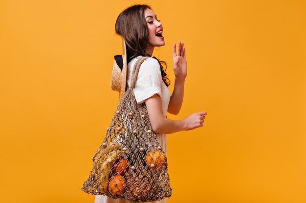 Charmante Frau im weißen Baumwollkleid wirft mit Stringtasche auf orange Hintergrund auf.