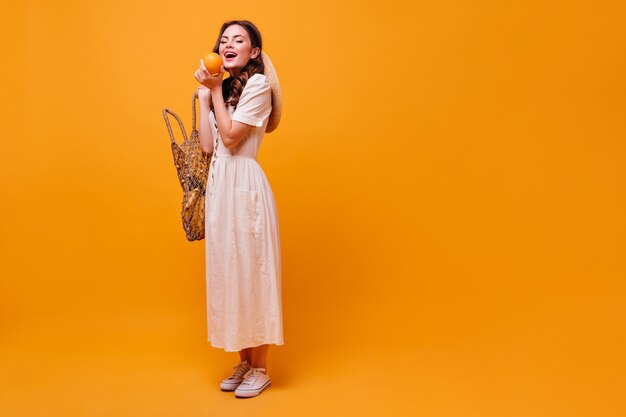 Charmante Frau im weißen Sommerkleid, das String-Tasche hält und Orange beißt.