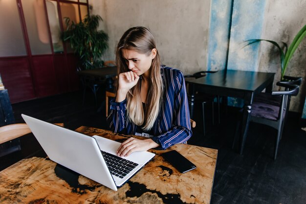 Charmante Frau in der blauen Jacke, die Computerbildschirm betrachtet. Innenfoto der langhaarigen Studentin, die im Café studiert.