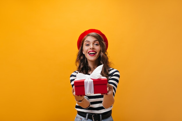 Charmante Frau in guter Laune hält rote Box auf orange Hintergrund. Attraktives Mädchen mit hellen Lippen im gestreiften Hemd freut sich über Geschenk.