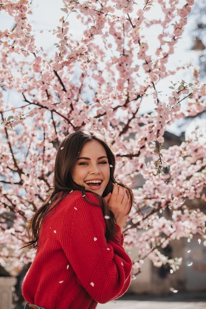 Charmante Frau in hellem Pullover lacht gegen blühende Sakura. Kühle brünette Frau im roten Outfit, das lächelt und Frühling genießt