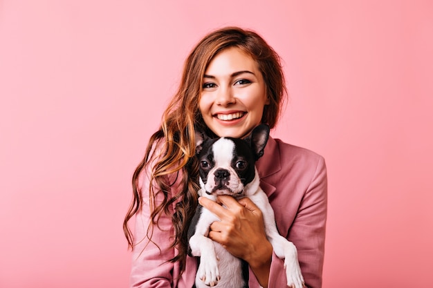 Charmante Frau mit glänzendem Ingwerhaar, das mit ihrem Haustier aufwirft. Gut gelauntes Mädchen in der rosa Jacke, die kleinen Hund hält.