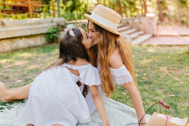 Charmante Frau mit lockigen langen Haaren lächelnd, während ihre Tochter sie küsst. Außenporträt des niedlichen kleinen Mädchens, das Spaß mit Mutter im Park mit Steinstufen hat.