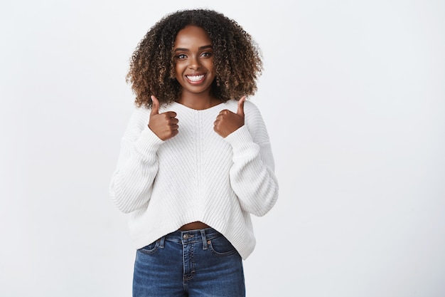 Charmante freundliche afroamerikanische frau mit lockigen haaren zeigt daumen hoch wie zustimmungsgeste, die im allgemeinen lächelt, stimme netten bedingungen zu, sage gute arbeit, stolzer freund hat das beste getan