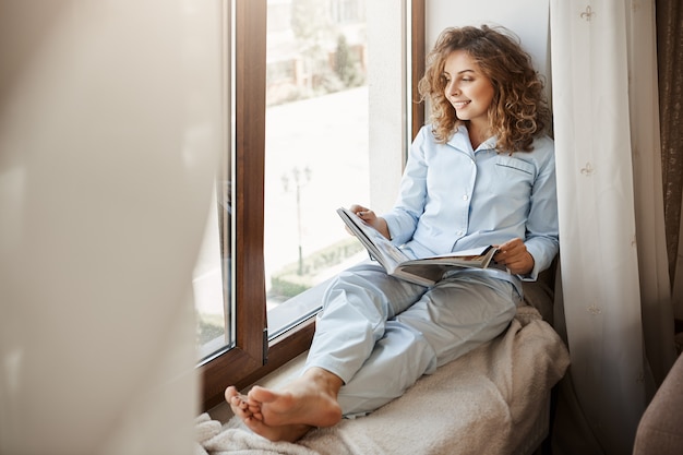 Charmante Geschäftsfrau, die entspannende Zeit zu Hause hat. Erfreute gut aussehende erwachsene Frau in Nachtwäsche, die auf dem Fensterbrett sitzt und auf die Straße schaut, Modemagazin hält und über Lebensstil liest