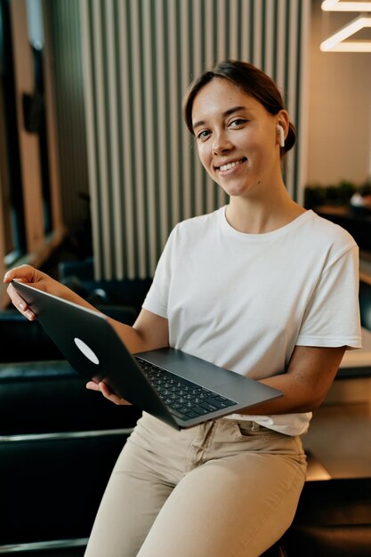 Charmante hübsche dame mit wundervollem lächeln und dunkel gesammeltem haar, gekleidet in weißes t-shirt und beige hose, arbeitet im büro mit laptop