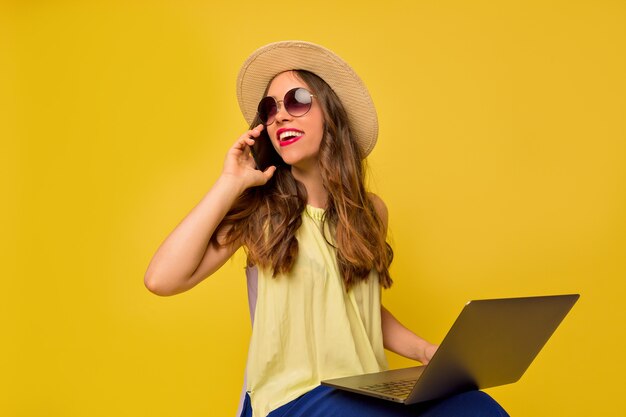 Charmante hübsche Frau mit Sommerhut und Brille, die am Telefon spricht und mit Laptop arbeitet