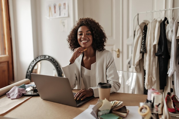 Charmante junge dunkelhäutige Frau in stylischer Jacke und Bluse lächelt Kamera arbeitet im Laptop und posiert im Büro