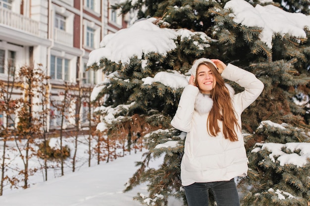 Charmante junge Frau, die an einem sonnigen, gefrorenen Morgen auf der Straße voller Schnee chillt. Fröhliches lächelndes Mädchen in warmer Kleidung, das Spaß auf Tannenbaumhintergrund hat. Glück, Freude, Positivität ausdrücken